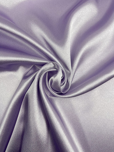 A close-up image of the smooth, shiny Satin Back Crepe - Lilac - 150cm fabric from Super Cheap Fabrics gathered in a swirl pattern. The lighting highlights the texture and sheen of the material, creating a sophisticated and elegant appearance.