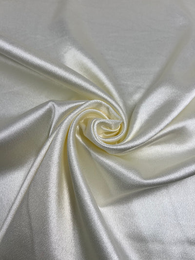 A close-up view of a piece of Super Cheap Fabrics' Satin Back Crepe - Ivory - 150cm fabric, elegantly twisted to create a soft, spiral pattern. The fabric has a shiny, reflective surface that highlights its luxurious texture and sheen, perfect for eveningwear.
