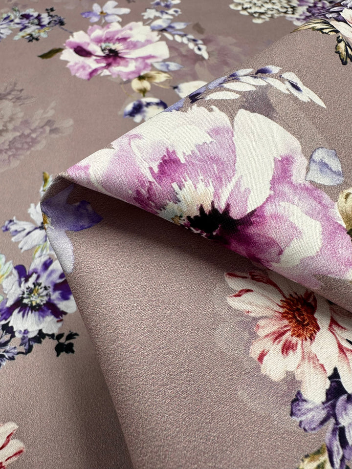 Close-up view of a Cotton Sateen - Shadow Rose - 120cm by Super Cheap Fabrics featuring a floral pattern with pink, purple, and white flowers on a light mauve background. The fabric is slightly folded, showcasing the detailed, vibrant multi-colour flower prints and the soft texture of the material.