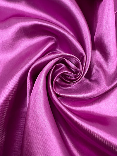 Close-up of shiny, pink Satin - Raspberry rose - 150cm fabric from Super Cheap Fabrics arranged in a swirling pattern, showcasing its smooth texture and reflective surface. The folds create a dynamic, spiral design, adding depth and movement to the image, perfect for glamorous satin dresses or elegant home décor accents.