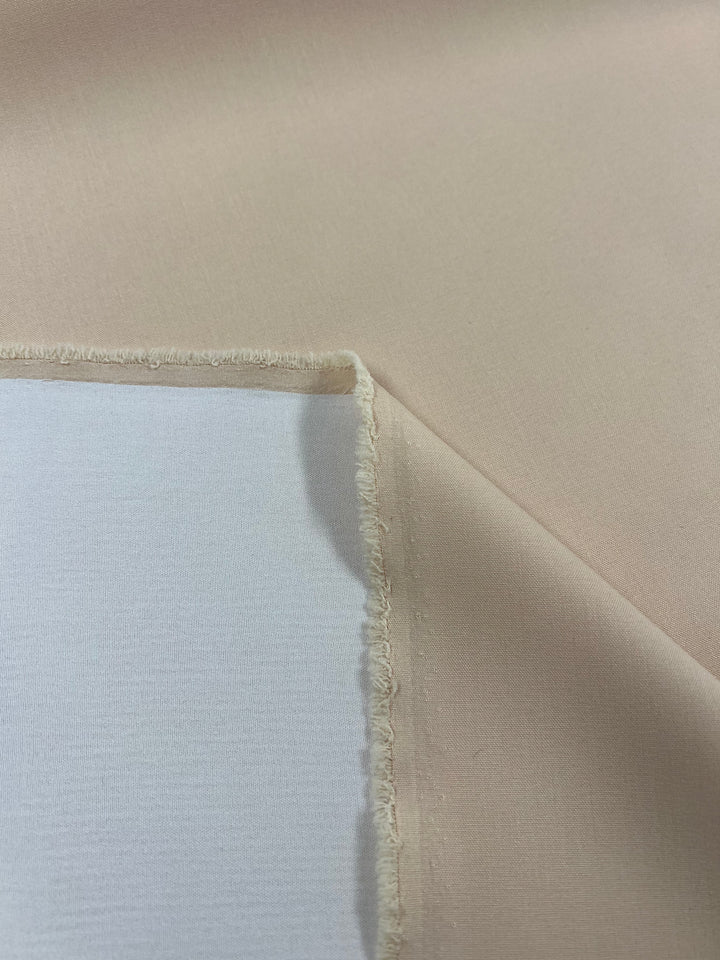 Close-up of a piece of beige Suiting - Amberlight - 145cm from Super Cheap Fabrics with a rectangular patch of white fabric on top. The edges have visible stitching, and the beige fabric, made from versatile polyester, is slightly folded, creating shadows and highlights.
