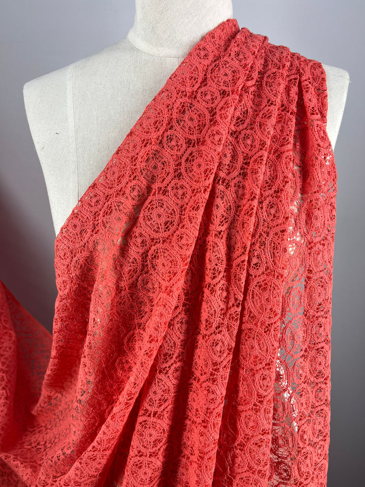 A mannequin draped with Super Cheap Fabrics' Lace - Georgia Peach - 150cm. The intricate lace pattern features circular motifs, creating an elegant and delicate appearance. The lightweight fabric is shown cascading over one shoulder, displaying its detailed design and texture.