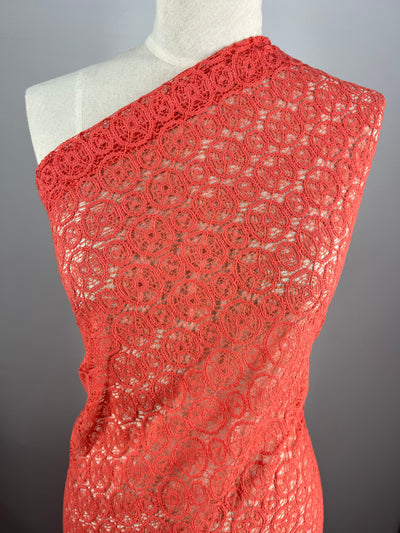 A mannequin draped in a red, intricately patterned lace fabric from Super Cheap Fabrics. The lightweight fabric, Lace - Georgia Peach - 150cm, covers the torso diagonally from one shoulder, revealing its delicate and elegant circular lace design. The background is a plain neutral gray.