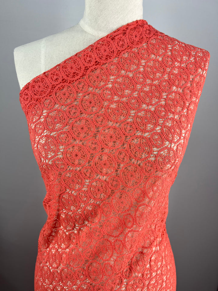 A mannequin draped in a red, intricately patterned lace fabric from Super Cheap Fabrics. The lightweight fabric, Lace - Georgia Peach - 150cm, covers the torso diagonally from one shoulder, revealing its delicate and elegant circular lace design. The background is a plain neutral gray.