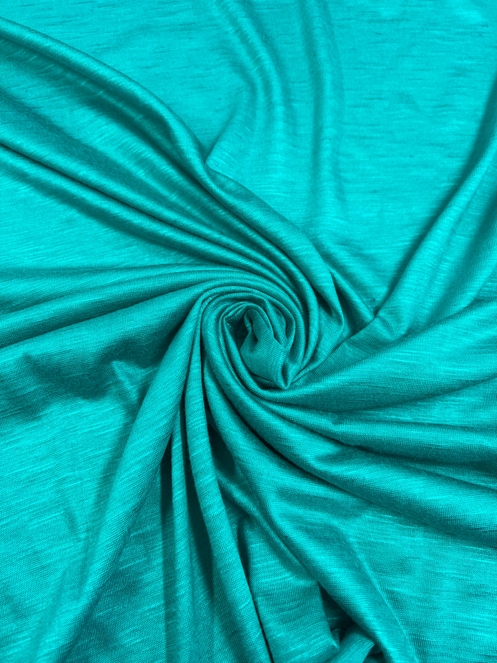 A close-up image of teal-colored Bamboo Jersey - Columbia - 170cm from Super Cheap Fabrics arranged in swirling folds, creating a circular pattern at the center. The fabric has a smooth texture, and the light highlights gentle wrinkles and shadows, enhancing the depth and movement of this environmentally responsible material.
