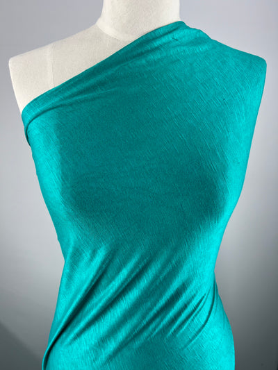 A mannequin draped in a piece of Bamboo Jersey - Columbia - 170cm from Super Cheap Fabrics, wrapped diagonally across the top and leaving one shoulder uncovered. The fabric is smooth, with a subtle sheen and soft texture. The backdrop is plain and grey, emphasizing the environmentally responsible choice of material.