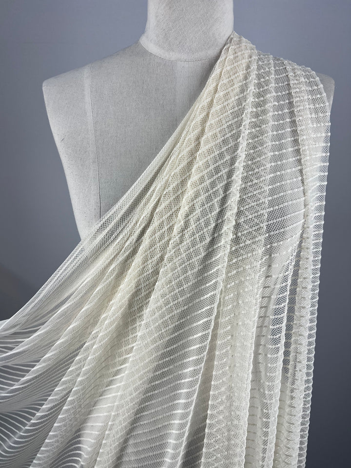 Close-up of a dress form draped with Super Cheap Fabrics' Lace - Ivory Mesh - 145cm. The lightweight fabric showcases intricate horizontal stripes and diamond-shaped patterns, creating a delicate, elegant appearance reminiscent of classic lace. The neutral gray background highlights the details of the 100% Polyester material perfectly.