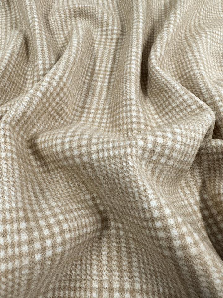 Close-up of a textured fabric featuring a beige and white checkered pattern. The heavyweight Wool Flannel - Macchiato - 148cm from Super Cheap Fabrics is draped in soft folds, creating gentle shadows and highlights that accentuate the checkered design, ideal for crafting cozy winter coats and jackets.