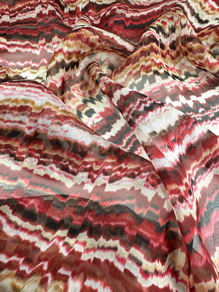 A flowing piece of luxury fabric with a vibrant, wavy pattern featuring streaks of red, brown, beige, and white colors. The Super Cheap Fabrics Pure Printed Silk - Celestial Warm - 140cm material has a textured appearance, showcasing a mix of sharp and blurred lines, creating a dynamic and visually striking design perfect for high-end fashion attire.