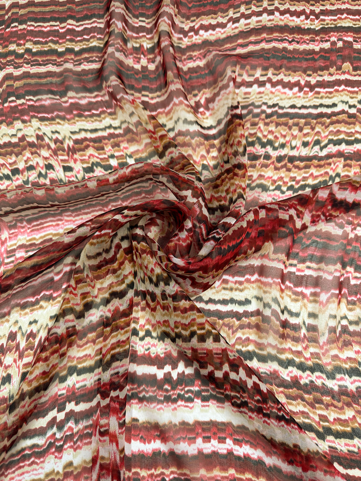 A close-up of Super Cheap Fabrics' Pure Printed Silk - Celestial Warm - 140cm with a swirling pattern of horizontal stripes in various shades of red, brown, and beige. The delicately crumpled center creates a dynamic and textured effect, perfect for high-end fashion attire.