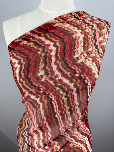A red, white, and black patterned Pure Printed Silk - Celestial Warm - 140cm fabric from Super Cheap Fabrics draped over a dress form. The luxury fabric features a wavy, abstract design with alternating colors and textures, creating a visually dynamic effect. The dress form partially covers the shoulder, showcasing the high-end fashion attire's intricate pattern.