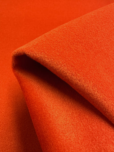 A close-up of a piece of **Super Cheap Fabrics' Wool Cashmere - Orange.com - 150cm** fabric draped and folded. The texture appears soft and slightly fuzzy, suggesting it could be made of a wool-cashmere blend or similar heavy weight fabric. The image captures the fabric's rich color and smooth, plush surface, ideal for luxurious outerwear coats.