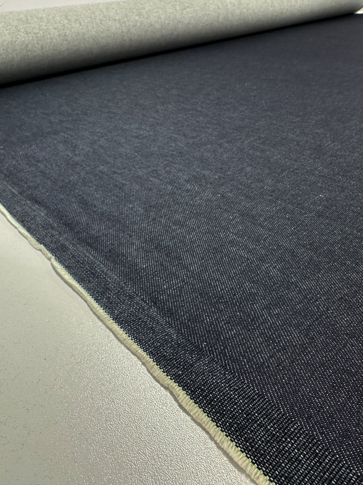 A close-up view of a large roll of 100% cotton, Designer Denim - Classic - 170cm by Super Cheap Fabrics, unrolled on a light grey surface. The texture and weave of the heavy weight denim are clearly visible, showing a fine, even pattern across the material. The edges of the fabric are neatly aligned.