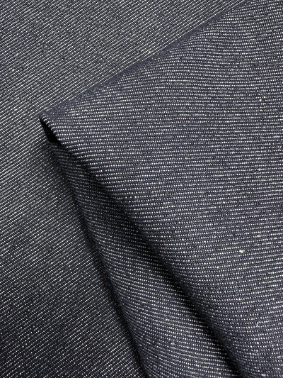 A close-up view of dark grey, heavy weight denim fabric with a subtle, fine diagonal weave pattern. The folded section of the 100% cotton material adds dimension and texture to the image. The Designer Denim - Classic - 170cm from Super Cheap Fabrics features small, intermittent specks that create a distinguished yet subtle visual effect.