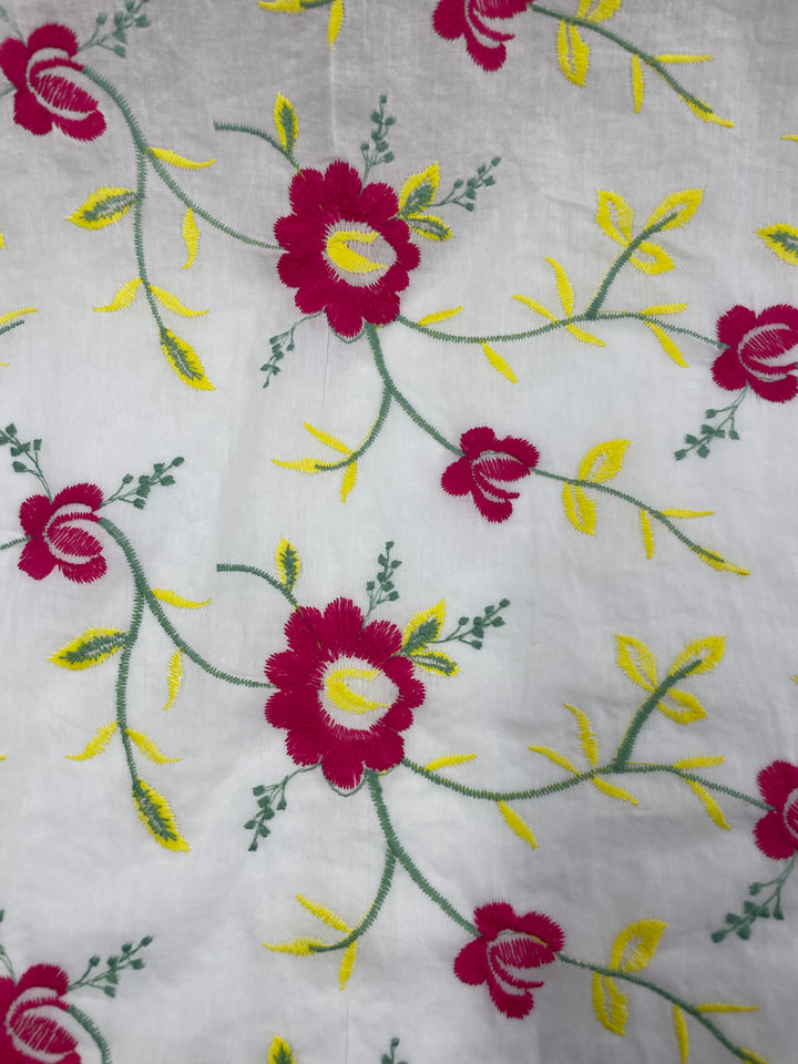 A close-up of Super Cheap Fabrics' Embroidered Cotton - Anemone - 150cm showcases a delicate floral pattern featuring red and yellow flowers, green leaves, and stems on a white background. The embroidery style is intricate with vibrant colors, capturing the beauty of this 100% cotton creation.