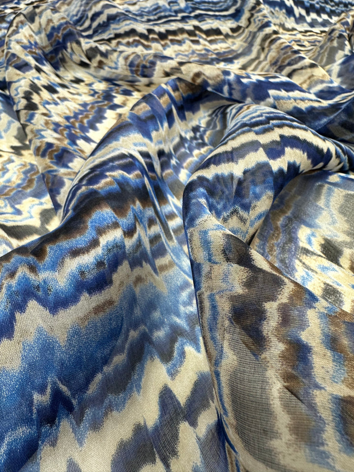 A close-up of Super Cheap Fabrics' Pure Printed Silk Chiffon - Celestial Cool - 135cm in a zigzag pattern featuring shades of blue, white, and black. This luxury fabric boasts a glossy, silky appearance, with a mix of light and dark tones creating a wavy, textured effect. The folds and ripples add depth to the intricate design, perfect for high-end fashion.