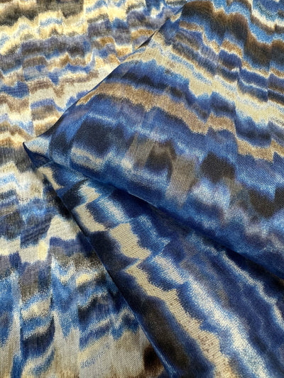 Close-up of a piece of luxury fabric with a wavy, abstract pattern in shades of blue, gray, and beige. The fabric appears to be Pure Printed Silk Chiffon - Celestial Cool - 135cm by Super Cheap Fabrics with a water-inspired design in darker and lighter hues, perfect for high-end fashion.