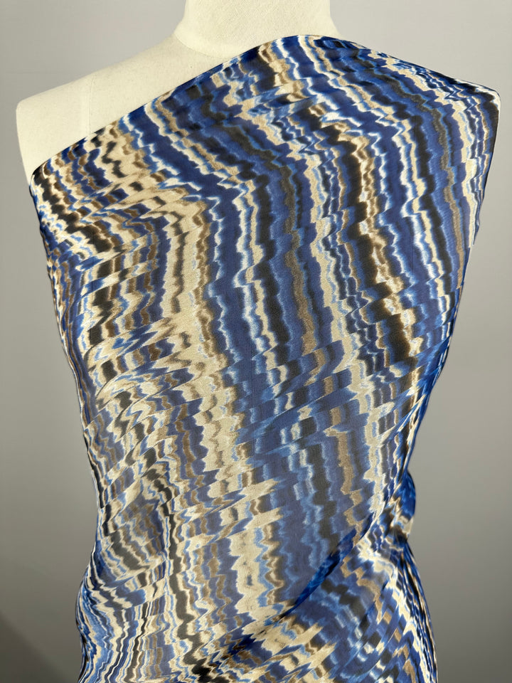 A draped Super Cheap Fabrics Pure Printed Silk Chiffon - Celestial Cool - 135cm fabric displayed on a dress form with an abstract, zigzag pattern in shades of blue, beige, and brown. The luxury fabric covers one shoulder and flows diagonally across the torso, creating an elegant and asymmetrical look for high-end fashion.