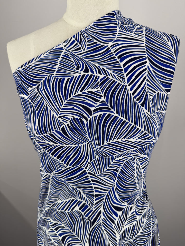 A mannequin is draped with Super Cheap Fabrics' Printed Lycra in Sapphire, featuring a bold blue and white abstract leaf design crafted from medium-weight Polyester/Spandex. Styled over one shoulder, it presents an elegant and modern look.