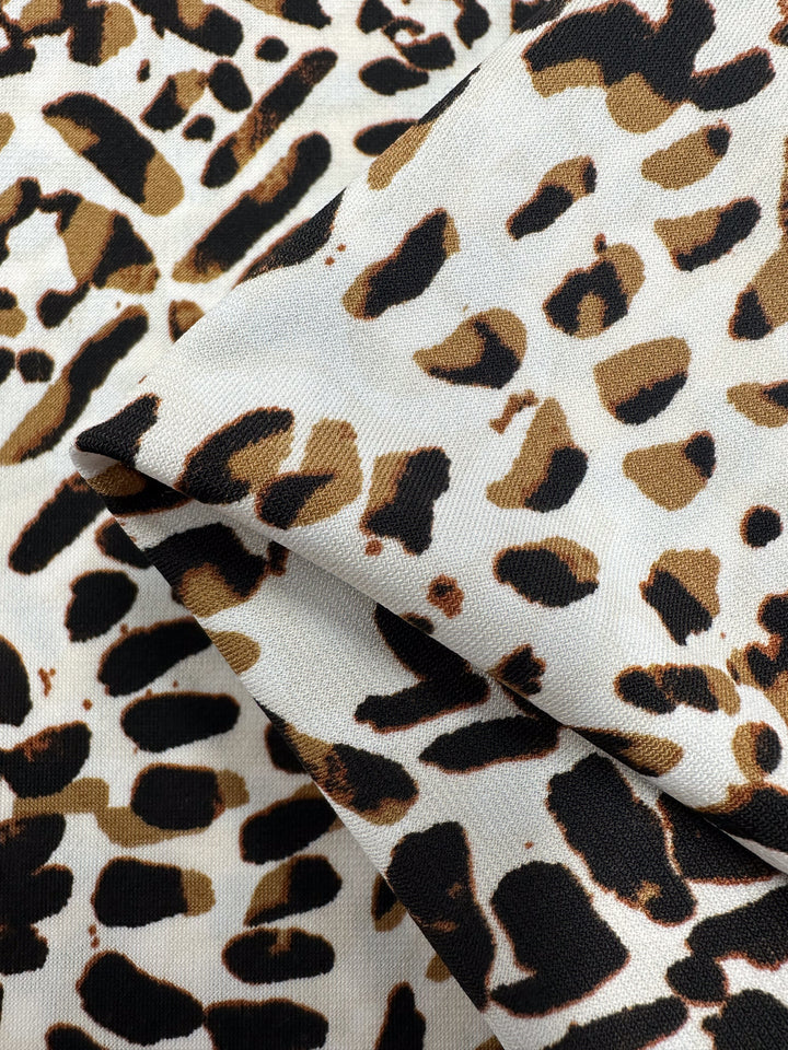 Close-up of a medium-weight fabric with a leopard print pattern. The Printed Lycra - Tanner - 150cm by Super Cheap Fabrics features spots in shades of brown and black on a beige background. The material appears soft and is slightly folded, showcasing the texture and detailed print.