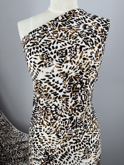 A mannequin draped in a one-shoulder dress made of medium-weight, Printed Lycra - Tanner - 150cm by Super Cheap Fabrics featuring a black and brown abstract animal print on a white background. The backdrop is plain grey with a piece of matching fabric in the lower left corner.