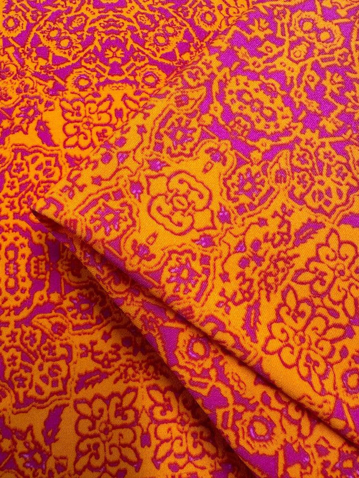 Brightly colored medium-weight fabric with an intricate floral and geometric pattern in bold shades of orange and magenta. The design features detailed flowers and abstract shapes, creating a vibrant and energetic visual effect. Several layers of Super Cheap Fabrics' Printed Lycra - Volcano Mosaic - 150cm are stacked together.