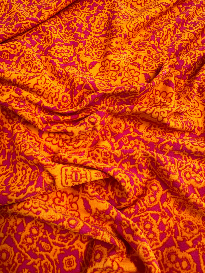 A close-up of a vibrant medium-weight fabric displaying an intricate pattern in shades of red, orange, and yellow. The design includes various abstract and floral elements, creating a bold, visually striking effect. This soft Printed Lycra - Volcano Mosaic - 150cm from Super Cheap Fabrics appears slightly wrinkled.