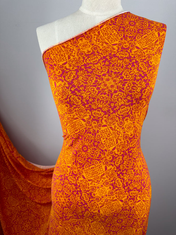 A draped mannequin displays a vibrant medium-weight fabric with an intricate orange and red pattern. The fabric cascades from the mannequin’s shoulder and down its side, showcasing the detailed design against a neutral gray background. The displayed product is Super Cheap Fabrics' Printed Lycra - Volcano Mosaic - 150cm.