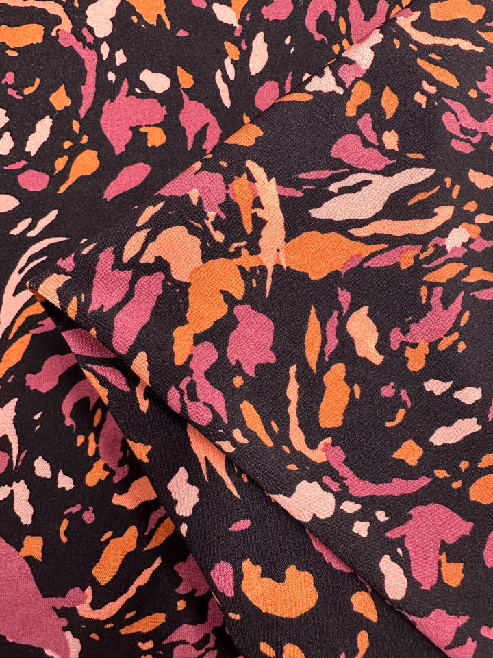 A piece of Super Cheap Fabrics Printed Lycra - Clawed - 150cm featuring an abstract pattern with swirling shapes in shades of pink, orange, and cream on a dark background. The fabric is folded, showcasing its vibrant design and soft texture.