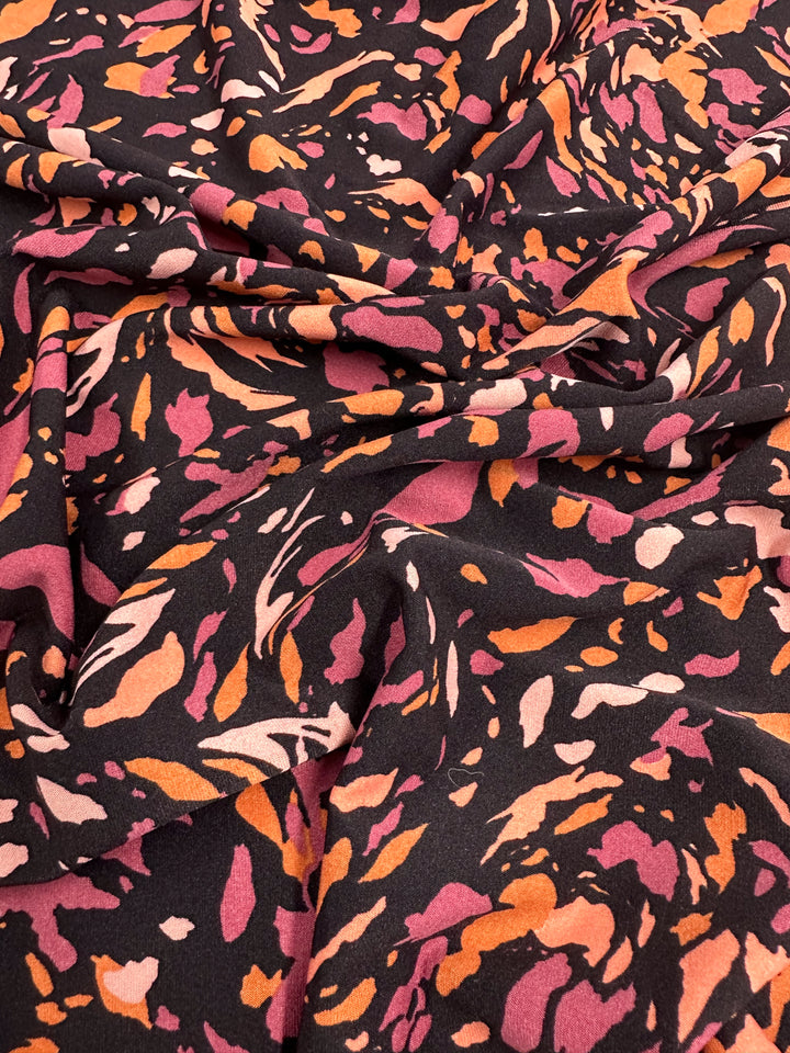 Close-up of a medium-weight fabric with a bold, abstract pattern. The design features irregular shapes and splashes in shades of pink, orange, and beige on a black background. The slightly draped Super Cheap Fabrics Printed Lycra - Clawed - 150cm creates soft folds and shadows.