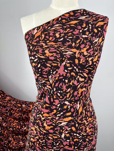 A close-up of a mannequin dressed in a one-shoulder dress made from Super Cheap Fabrics' Printed Lycra - Clawed - 150cm. The pattern consists of vibrant splashes of orange, pink, and beige on a black background. The dress is displayed against a plain, light-colored backdrop.