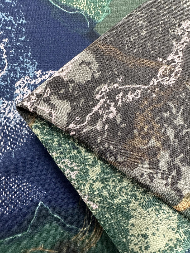 A close-up photograph showing two pieces of medium-weight fabric with camouflage patterns overlapping. One fabric, made of Polyester/Spandex, has shades of blue and white, while the other shows a mix of grey, green, and brown tones. The textures and stitching are clearly visible. This fabric is the Printed Lycra - Drifter - 150cm by Super Cheap Fabrics.
