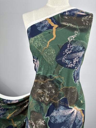 A mannequin is draped with a single-shoulder garment featuring a modern abstract pattern. The medium-weight fabric design includes shades of green, blue, gold, and white with organic, irregular shapes and textured details. The background is a plain gray. The fabric used is the Printed Lycra - Drifter - 150cm from Super Cheap Fabrics.