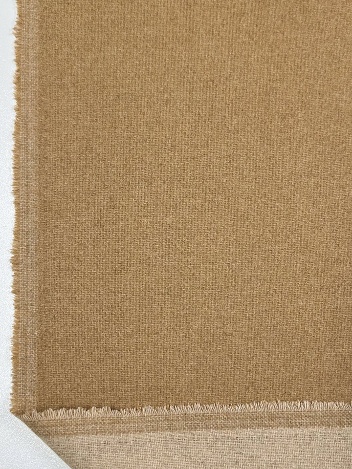 A close-up image of a piece of brown fabric showcases its slightly rough texture and frayed edges. Made of Reversible Merino Wool - Latte - 150cm by Super Cheap Fabrics, the fabric's top part is darker brown, while the underside appears lighter and smoother. The natural moisture-wicking properties make it ideal for comfortable wear.