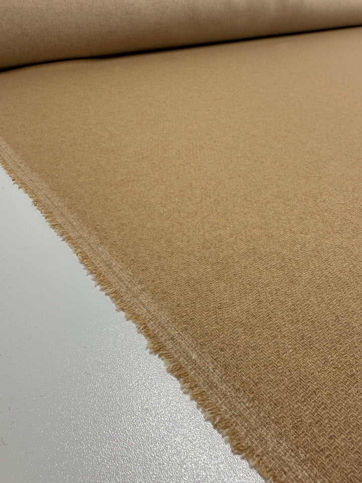 A roll of beige Reversible Merino Wool - Latte - 150cm from Super Cheap Fabrics with a rough, unfinished edge is spread out on a flat surface. The texture appears soft and woven, boasting excellent breathability. The image captures the material close-up from a low angle, emphasizing the details of the weave and edge.