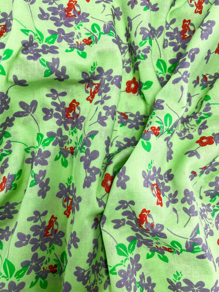 A close-up view of the Linen Voile - Jazzy - 140cm by Super Cheap Fabrics showcasing its vivid green fabric adorned with a floral pattern. The breathable material features purple flowers, small red blooms, and green leaves, creating a colorful design ideal for home decor. The fabric appears to be slightly wrinkled.