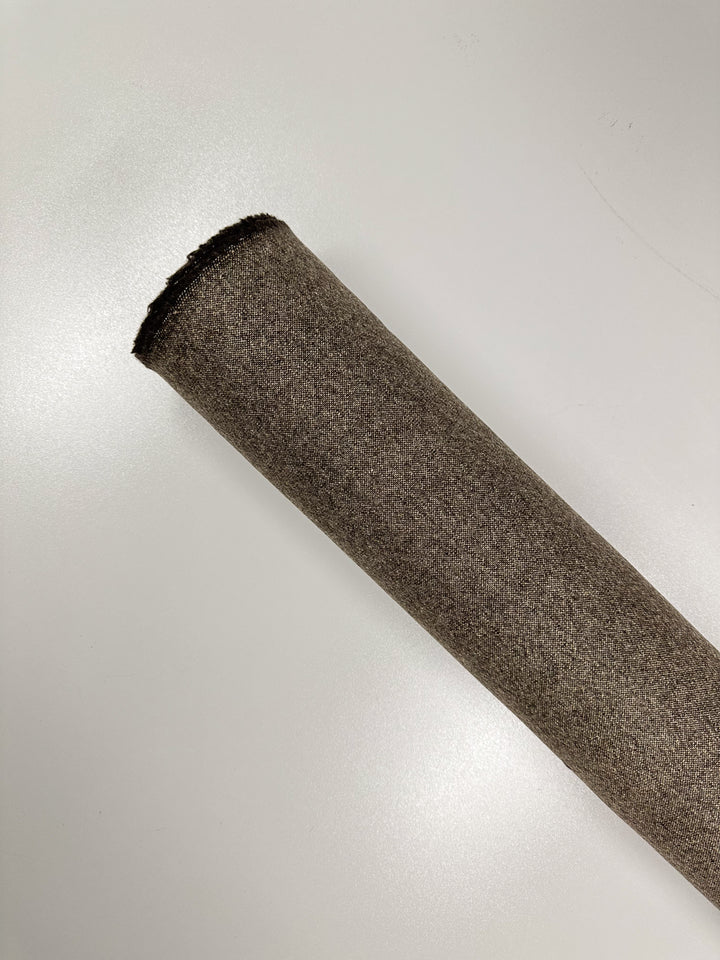 A rolled-up piece of medium weight wool fabric with a textured appearance, placed on a white surface. The roll, likely crafted in Italy from fine wool tweed, is partially angled to the left and seen from a top-down perspective. This is "Wool Tweed - Speckled Brass - 147cm" by Super Cheap Fabrics.