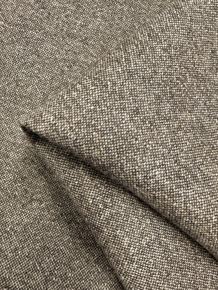Close-up image of a folded piece of Super Cheap Fabrics' Wool Tweed - Speckled Brass - 147cm with a coarse, textured appearance. The fabric is woven in a tweed-like pattern, featuring a mix of neutral tones including gray and taupe. The intricate interweaving creates a speckled effect reminiscent of classic Italian textiles.