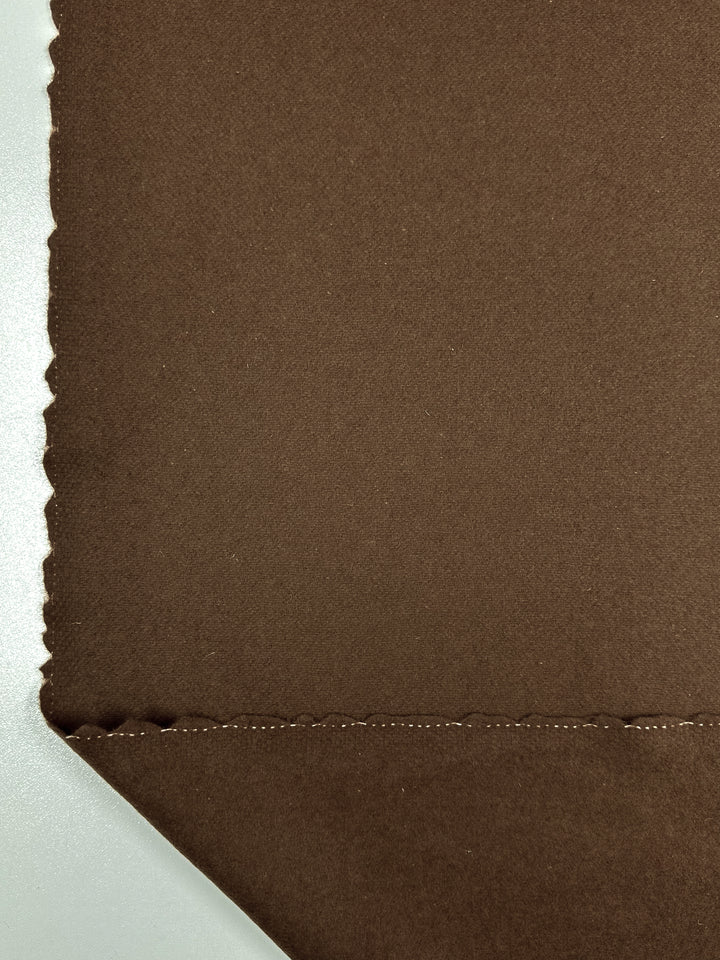 A close-up image of a brown Melton Wool - Caramel Cafe - 150cm fabric swatch by Super Cheap Fabrics with a neat diagonal cut at the bottom corner. The right edge of the swatch has a scalloped pattern, while the bottom has a straight seam with visible stitching. The background is a solid light color.