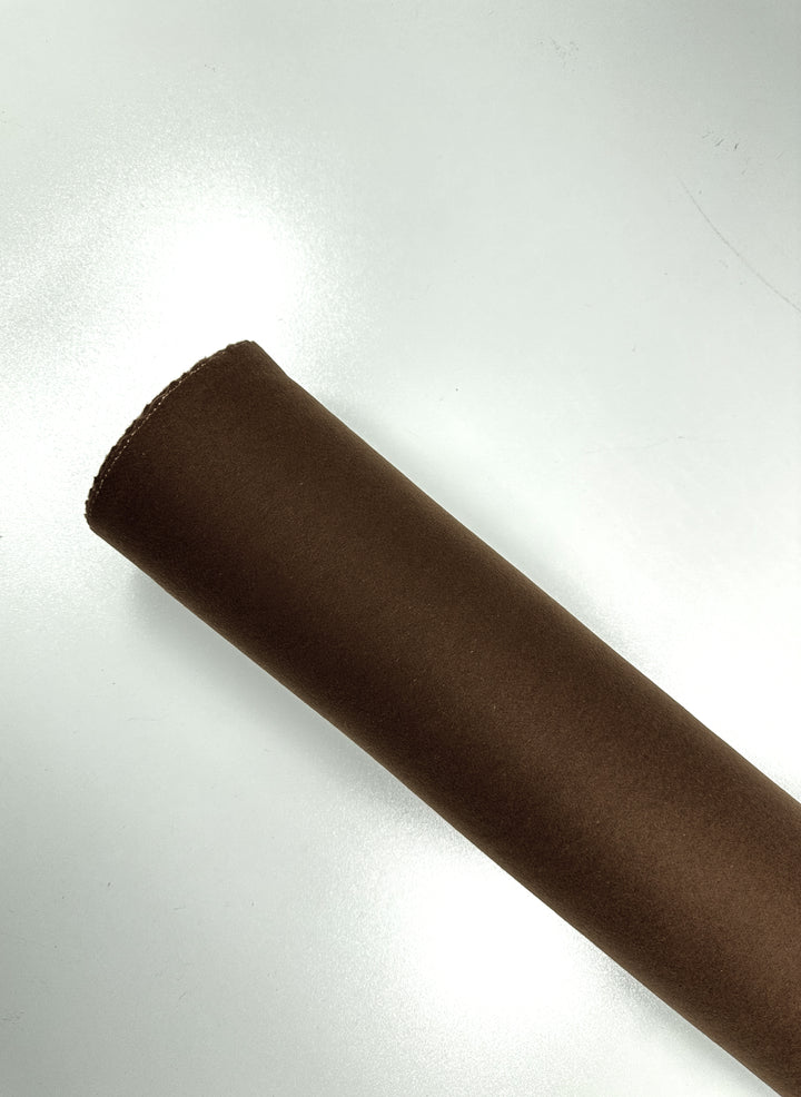 A roll of Super Cheap Fabrics' Melton Wool - Caramel Cafe - 150cm is positioned diagonally from the bottom left to the top right against a plain, light-colored background. The heavy weight wool fabric appears to have a smooth, matte texture.