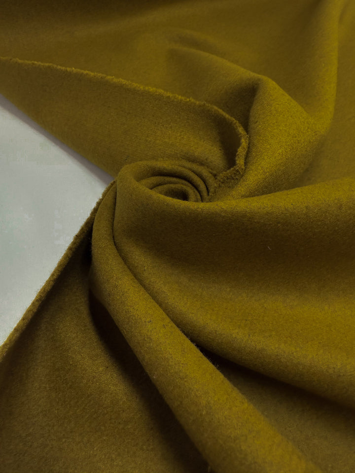 A close-up of Super Cheap Fabrics' Wool Cashmere - Plantation (155cm) in mustard yellow, arranged in soft folds and a swirl pattern to highlight its smooth, slightly textured surface.