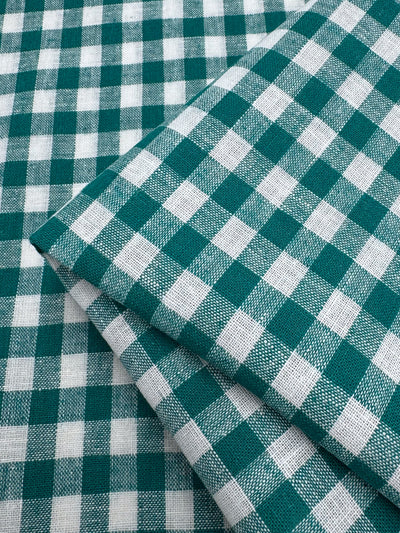 A close-up of Super Cheap Fabrics' Linen Blend - Verdant Gingham (145cm) shows two lightweight pieces in a classic green and white check pattern, neatly folded on a matching background.