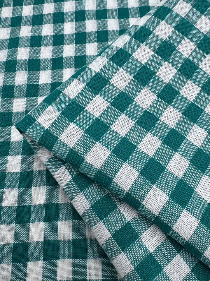 A close-up of Super Cheap Fabrics' Linen Blend - Verdant Gingham (145cm) shows two lightweight pieces in a classic green and white check pattern, neatly folded on a matching background.