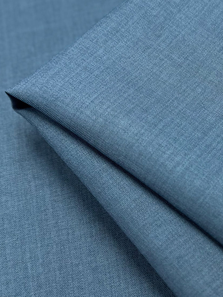 A close-up of Twill Suiting - Bluesteel by Super Cheap Fabrics shows neatly folded blue fabric with a smooth texture, fine diagonal weave, clean lines, and subtle shading.