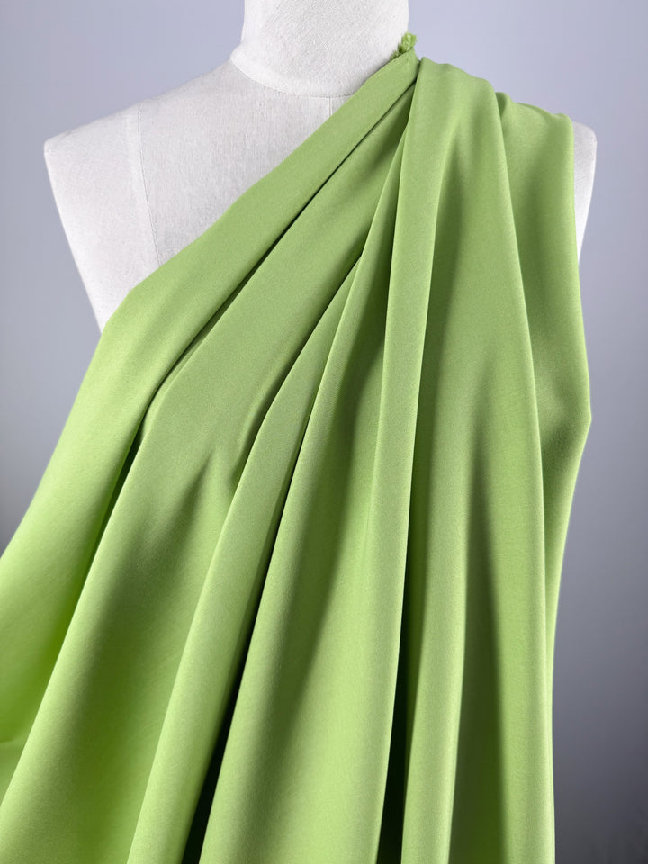 Super Cheap Fabrics’ Twill Suiting - Green Glow (150cm), a lime green polyester with a subtle diagonal weave, is shown draped in soft folds across one shoulder on a white dress form.