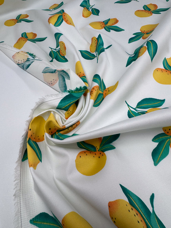 Super Cheap Fabrics' Cotton Sateen - Lemon Grove (145cm) features a white base with vibrant yellow lemons and green leaves, slightly gathered and twisted at the center to display the colorful fruit design, with visible fabric edges on the left.
