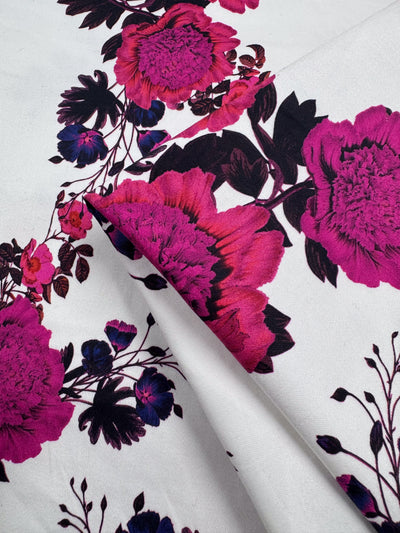 Super Cheap Fabrics’ Cotton Sateen - Bold Bloom (142cm) features a white medium-weight fabric with large bright pink flowers and dark purple leaves—ideal for household décor. Partially folded to highlight its vibrant design.