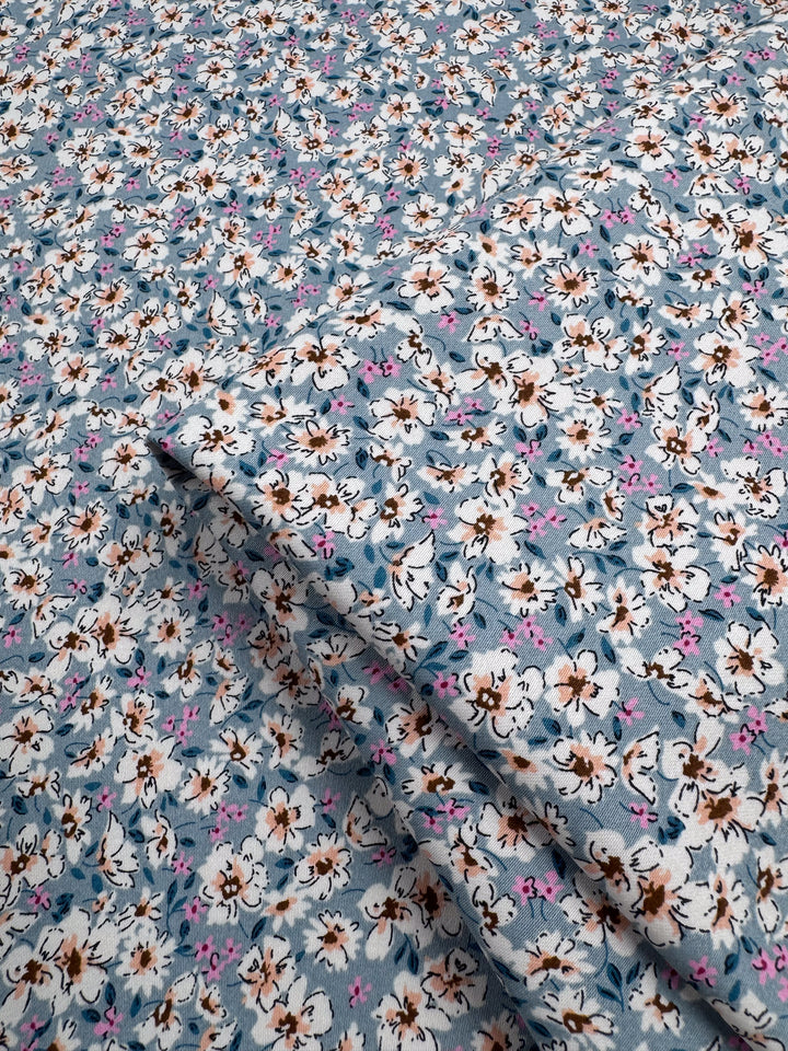 A close-up of Super Cheap Fabrics’ Cotton Sateen - Bluebell Meadows (150cm) shows neatly folded blue fabric with white, brown, and pink floral patterns—ideal for multi-use projects.