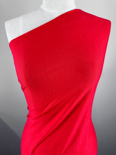 A mannequin is draped in a vibrant Bittersweet rayon lycra one-shoulder garment from Super Cheap Fabrics. The medium weight two-way stretch fabric has a soft texture and covers the torso in an elegant, flowing manner. The background is a gradient of grey and white, making the bright red fabric stand out prominently.
