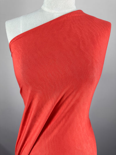 A mannequin draped in Super Cheap Fabrics Rayon Lycra - Hot Coral - 145cm fabric. The two-way stretch material is wrapped around the mannequin's torso with one shoulder exposed, creating a diagonal line across the chest. The background is a plain, light gray color.